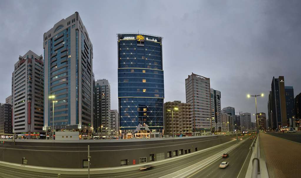 占奈萨拉卜塔酒店 阿布扎比 外观 照片 The photo depicts a modern urban landscape featuring several high-rise buildings. In the foreground, there is a road with vehicles, suggesting a busy city environment. Prominent among the buildings is a tall, blue structure with a distinctive logo at
